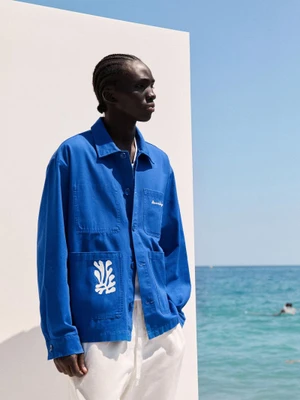Model in a blue jacket with white graphic detail standing against a white wall with the ocean in the background