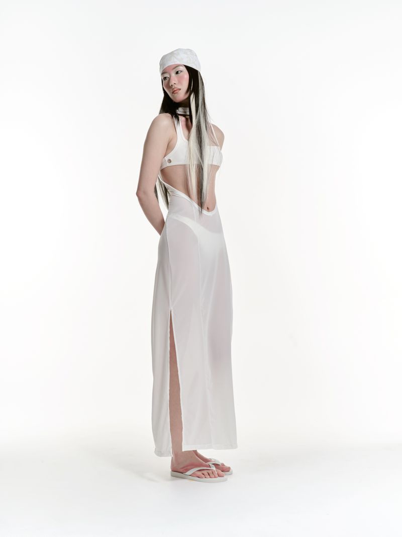 Full shot of a young woman with long dark hair, wearing a white headscarf, bikini top, and a sheer dress, standing in a bright white studio.