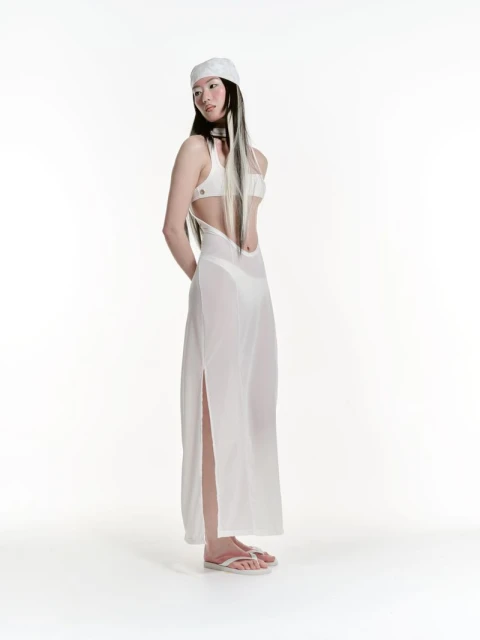 Full shot of a young woman with long dark hair, wearing a white headscarf, bikini top, and a sheer dress, standing in a bright white studio.
