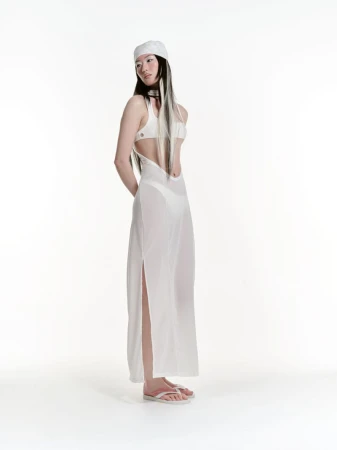 Full shot of a young woman with long dark hair, wearing a white headscarf, bikini top, and a sheer dress, standing in a bright white studio.