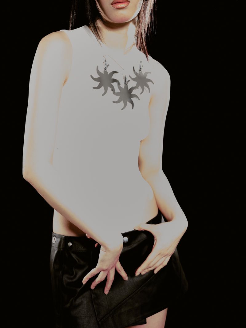 Cropped view of a model wearing a white graphic sleeveless top with sun motifs and a black low-rise mini skirt against a black background.