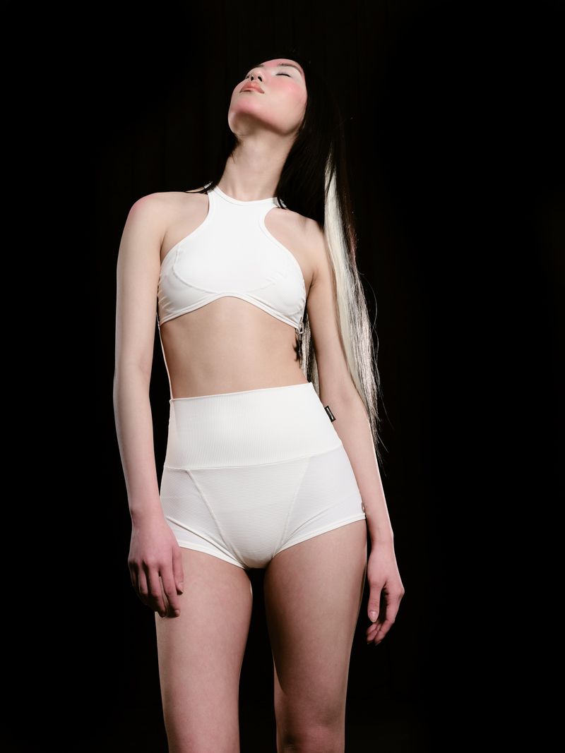 Asian woman with long black and blonde hair poses in a high-neck top and high-waisted shorts against a stark black backdrop.