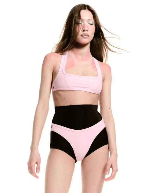 Brunette fashion model with closed eyes poses in pink bikini top and black shorts on white backdrop, minimalist fashion concept.