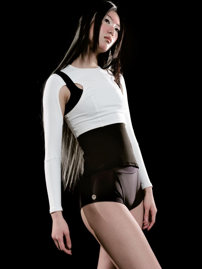 Athletic-looking Asian woman posing in white long-sleeved top with black shorts and waistband against a dark backdrop.