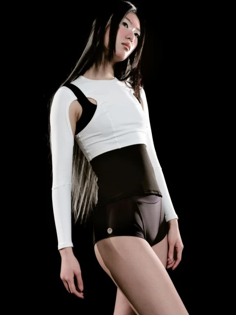 Athletic-looking Asian woman posing in white long-sleeved top with black shorts and waistband against a dark backdrop.