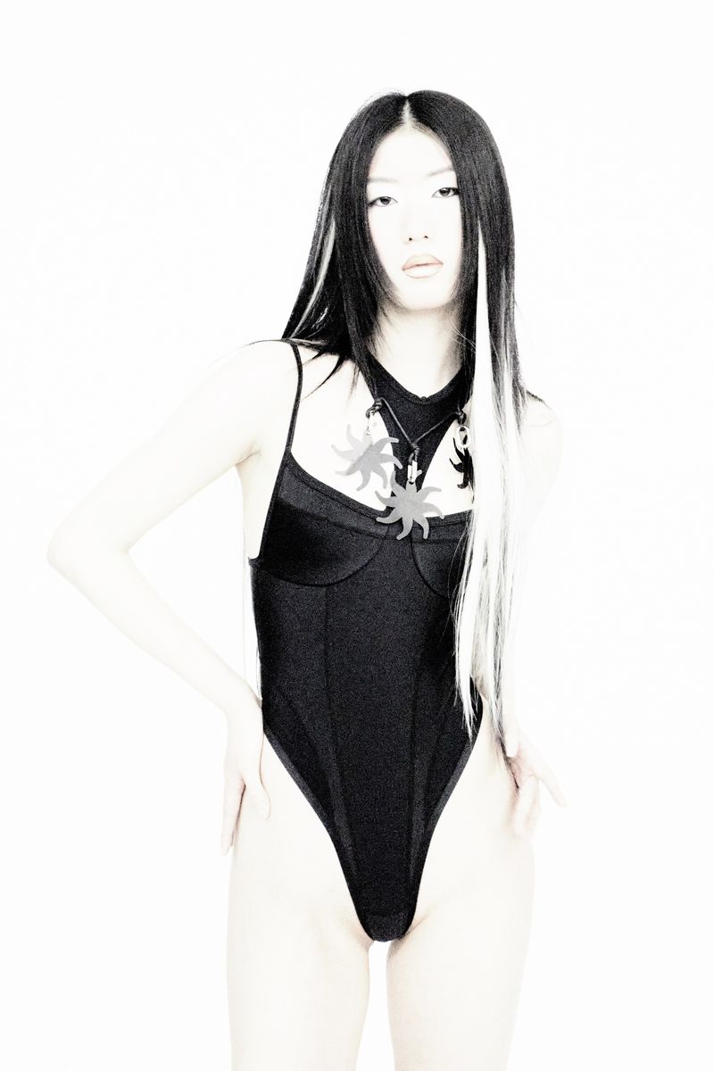 Asian woman in black bodysuit with sun pendants on a white backdrop, hands on hips, showcasing fashion and style with long hair.
