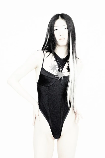 Asian woman in black bodysuit with sun pendants on a white backdrop, hands on hips, showcasing fashion and style with long hair.