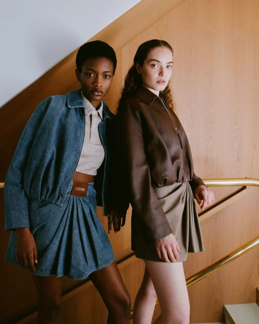 Two women in fashionable denim and leather jacket outfits pose in an indoor stairwell, showcasing modern style and design.