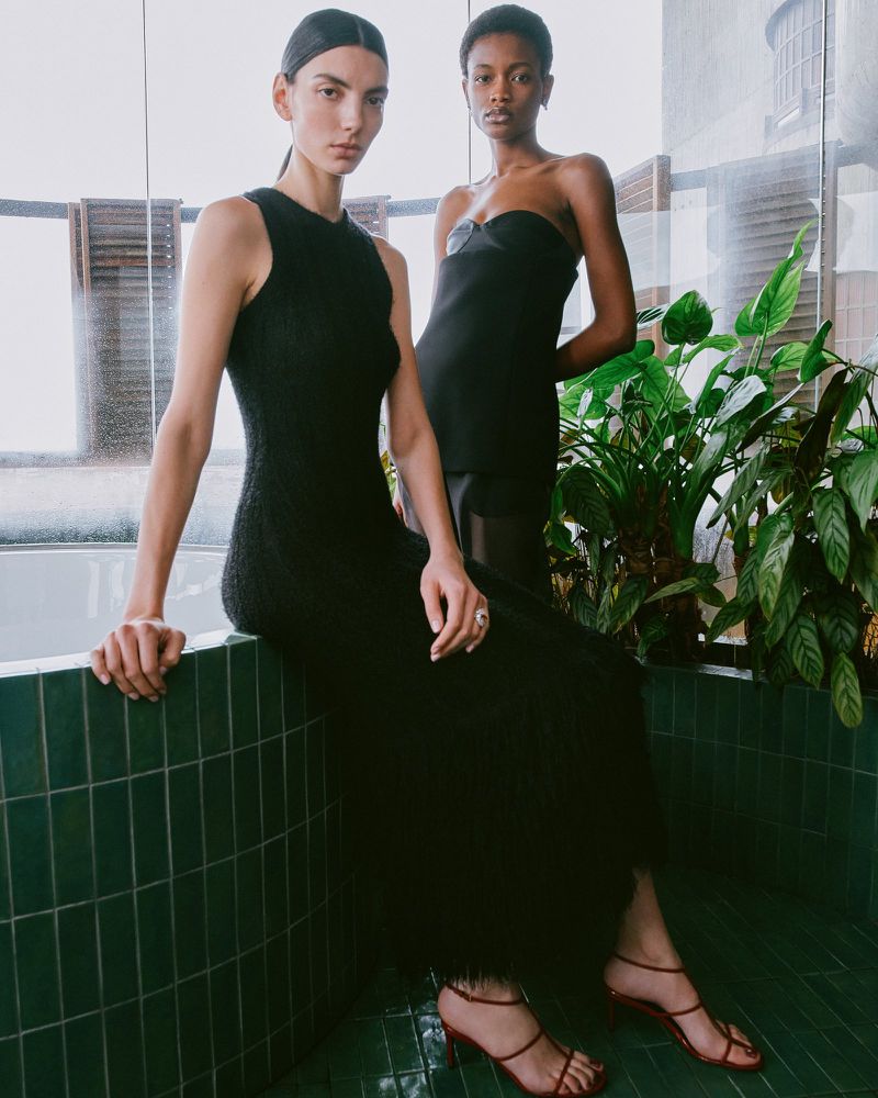 Two women pose indoors wearing elegant black dresses and strappy red heels, with natural light and lush green plants in the background.