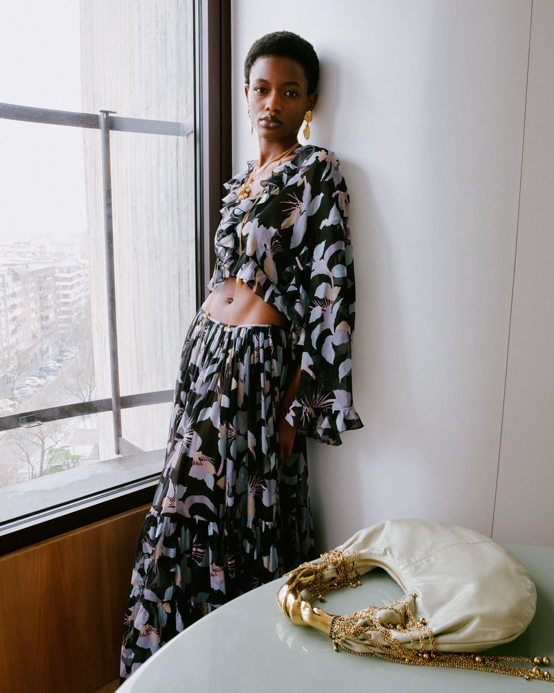 Fashionable woman in floral two-piece outfit poses by window next to designer handbag. Building visible through the window.