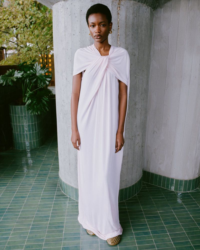 An African-American woman in a light pink gown poses indoors next to a fluted concrete pillar and green tile flooring.