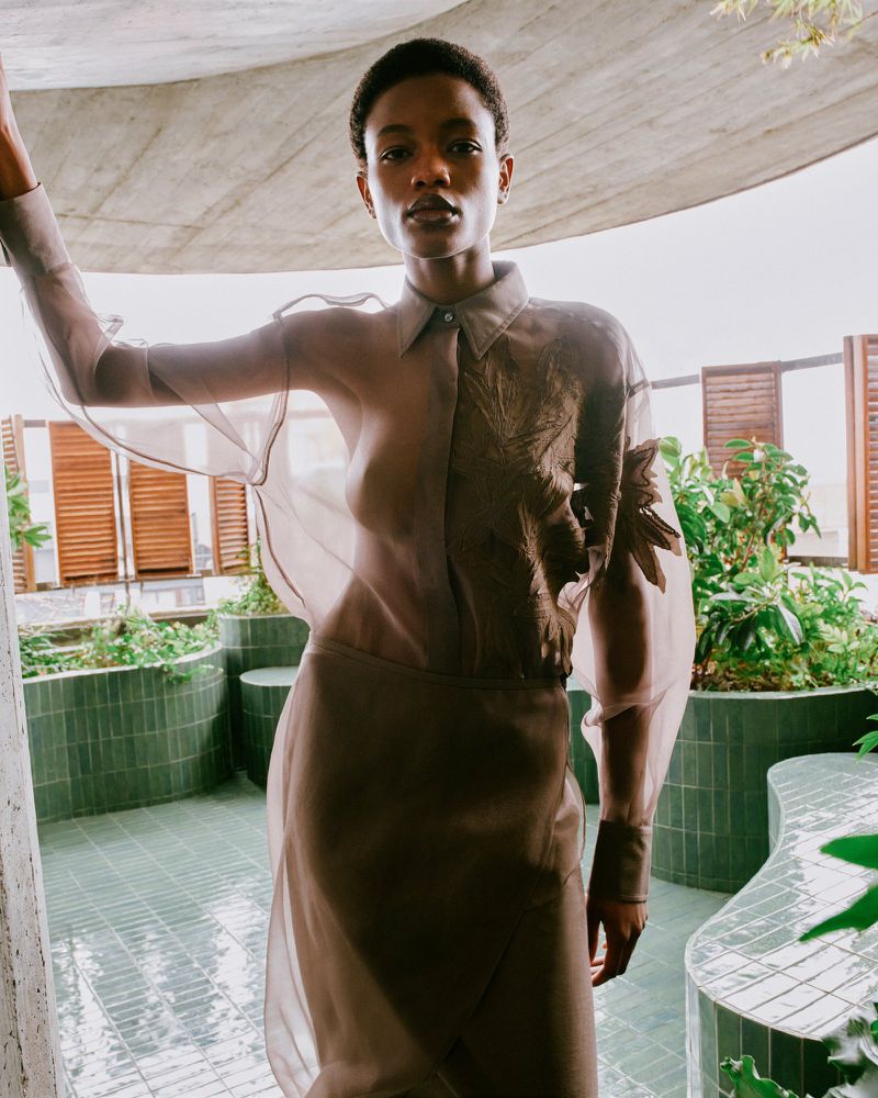 Fashion shot of a Black woman in a sheer, collared blouse with a brown skirt, posing on a tiled balcony with plants and dappled light.