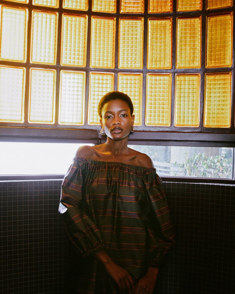 Portrait of a slender African-American woman in an off-the-shoulder dress, posing in front of a window with patterned glass blocks.