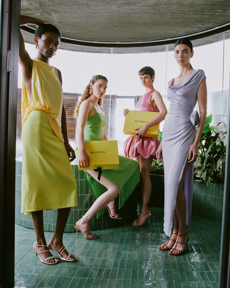 Four fashion models pose indoors wearing dresses in yellow, green, pink, and lavender, holding yellow boxes from MYTHERESA in a studio setting.