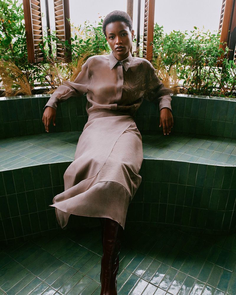 A Black woman poses seated on a tile bench, wearing a button-up shirt, knee-length skirt, and knee-high boots in a neutral indoor setting.