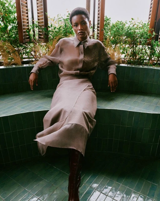 A Black woman poses seated on a tile bench, wearing a button-up shirt, knee-length skirt, and knee-high boots in a neutral indoor setting.