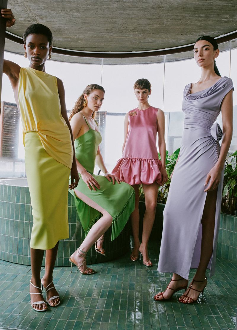 Four women pose in pastel-colored dresses and high-heeled sandals, standing indoors on a green tiled floor. Fashion photography, full shot.