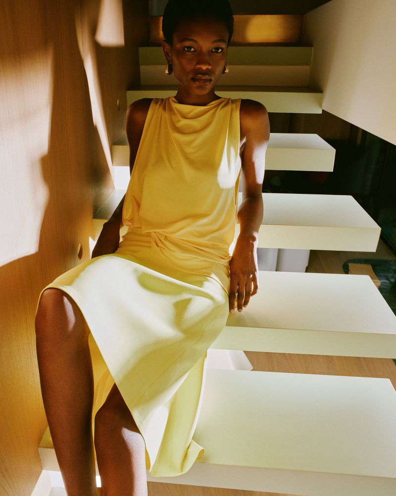 Portrait of an adult female model in a light yellow dress sitting on white stairs, with soft lighting and a serene expression.