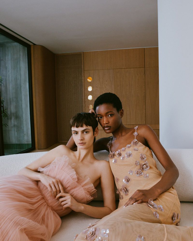 Two women pose on a white couch in a neutral room wearing formal dresses, one blush tulle and one nude with floral embellishments.