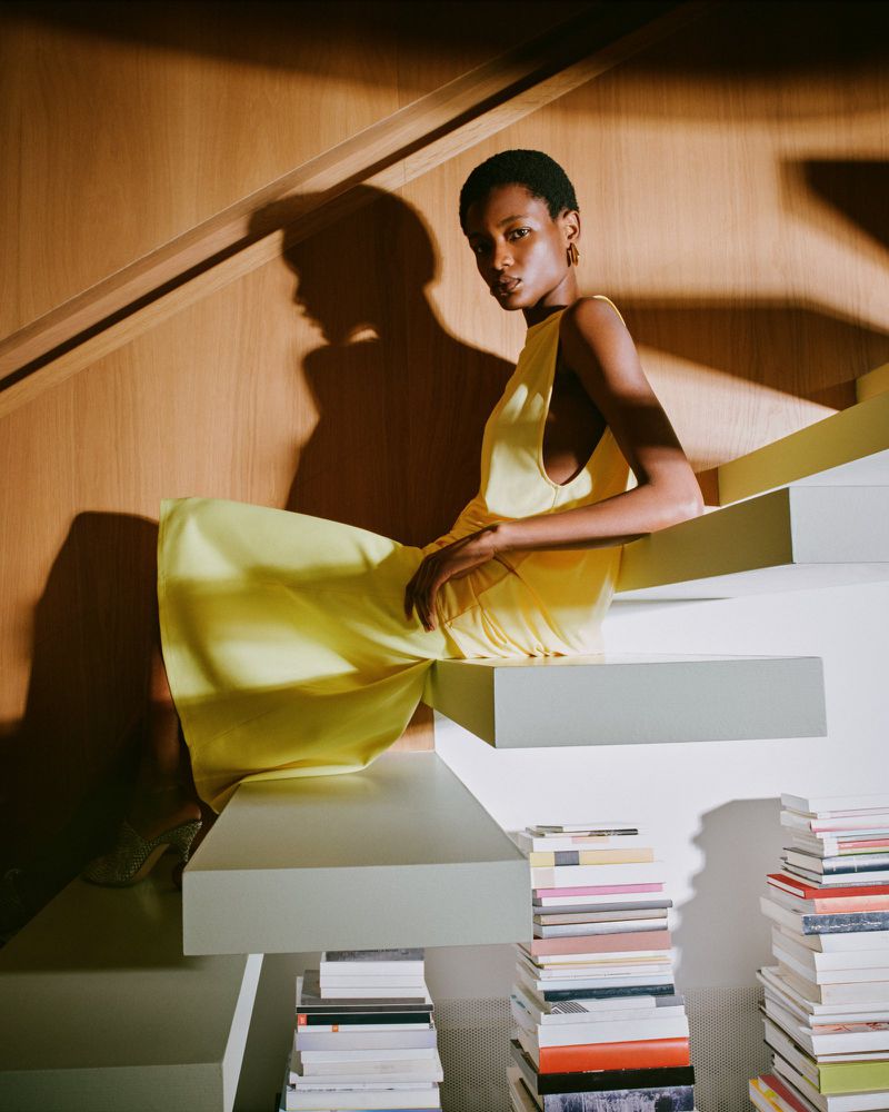 Elegant woman in a yellow dress poses on white steps near a stack of books, accented by the shadows and light from a staircase.