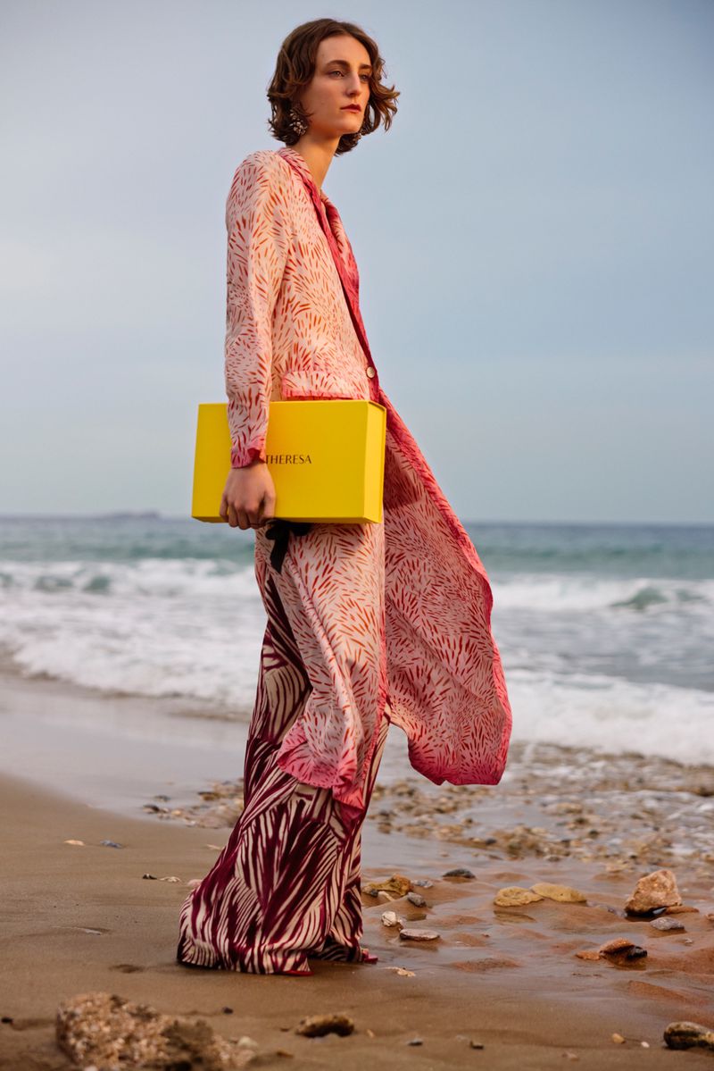 Woman with a yellow box stands on a sandy beach wearing a patterned pink jacket and pants in an ocean landscape setting with rocks.