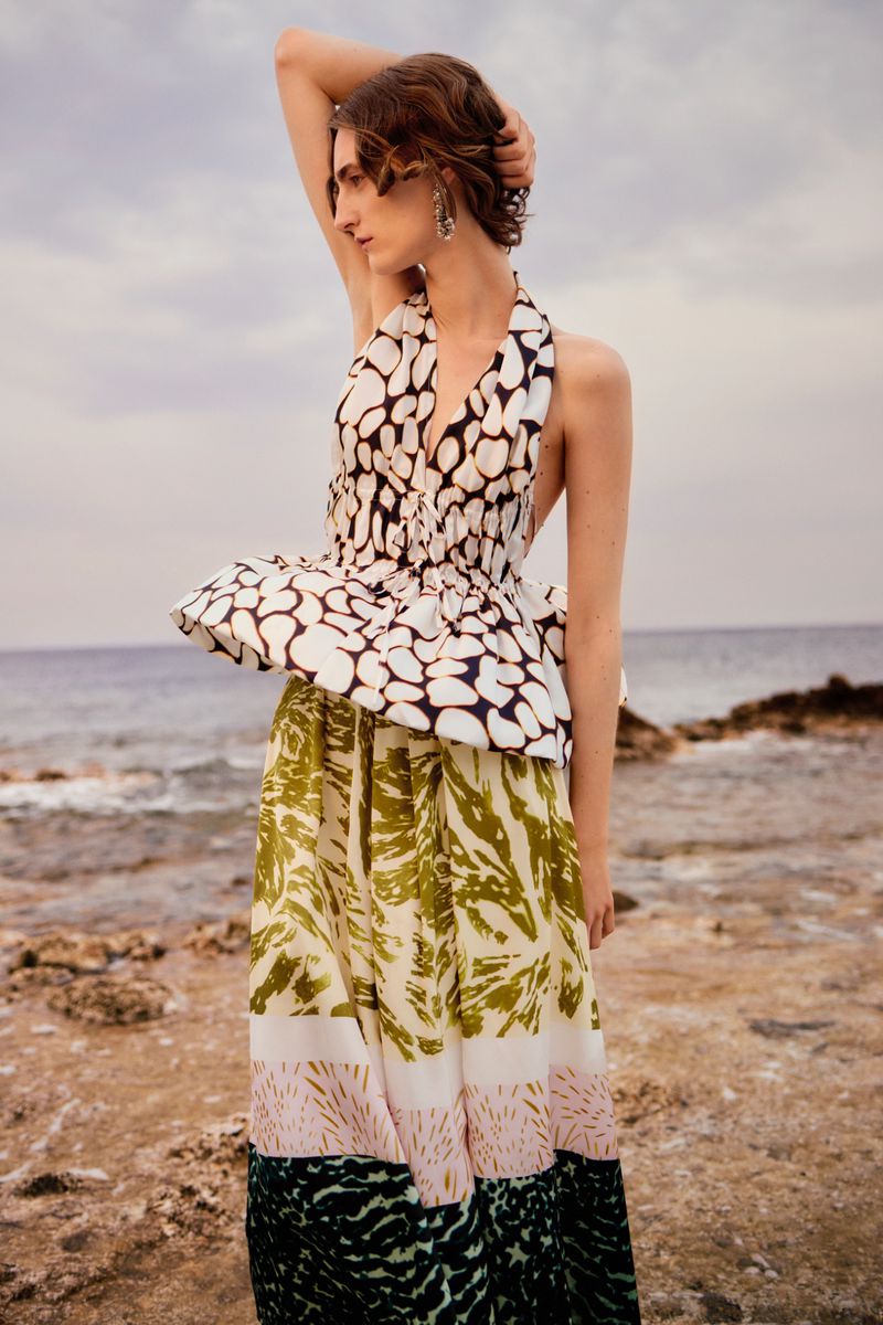 Elegant fashion shot: a brunette woman poses on a beach, wearing a stylish halter top and a long skirt with olive and black botanical prints.