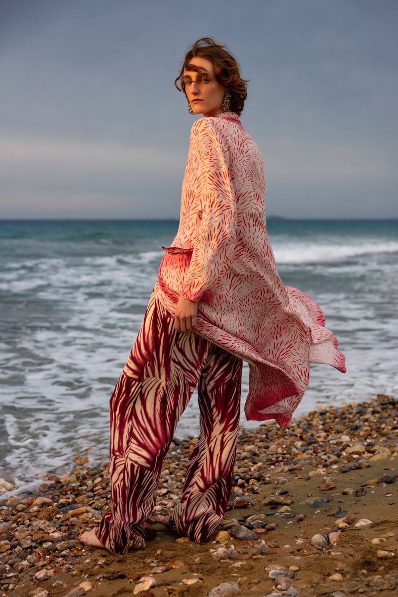Young woman in patterned robe and pants on rocky beach with ocean backdrop; fashion shoot, outdoor setting, overcast lighting.