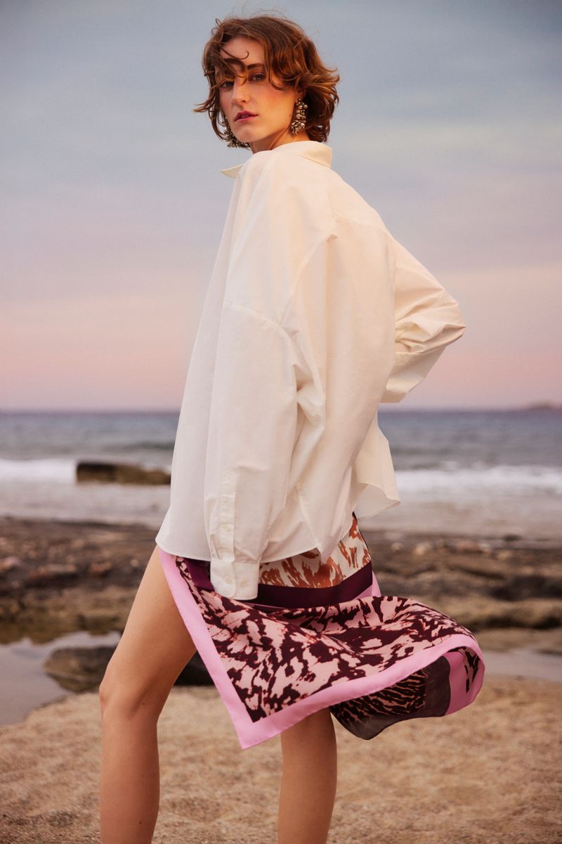 Woman posing in white shirt and patterned skirt on rocky beach at sunset. Fashion shot with ocean and sky backdrop.