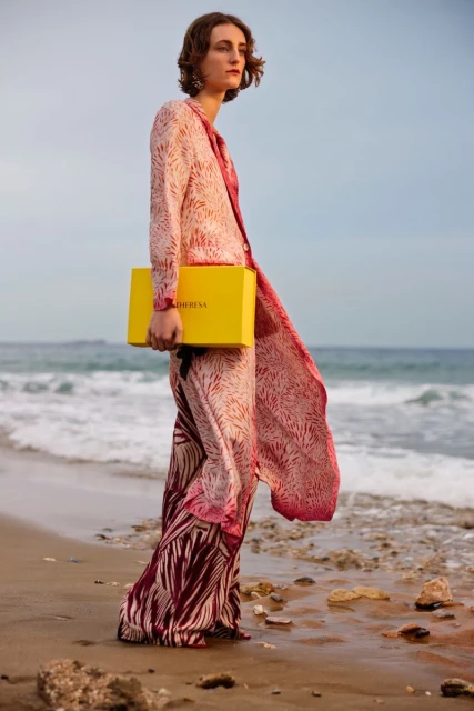 Model Ines Pottier in a patterned pink coat holding a bright yellow Mytheresa box while standing on a sandy ocean beach.