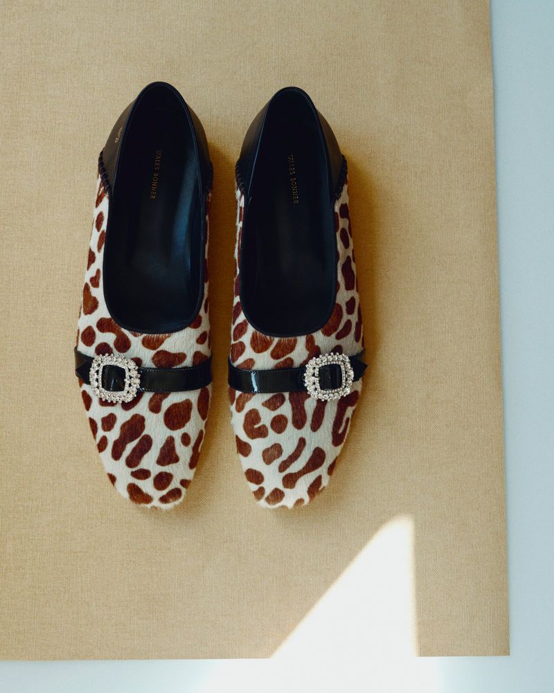 Overhead studio shot of a pair of ballet flat shoes with a brown-and-white spotted print and black patent leather trim and buckle.