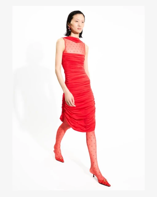 A full shot of a slender model walking in a red, ruched dress with matching tights and heels against a white background in a fashion editorial.