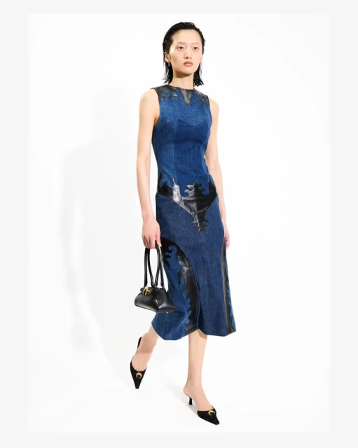 Elegant woman in a blue denim dress and black heels stands in a white photo studio holding a black purse, fashion forward.