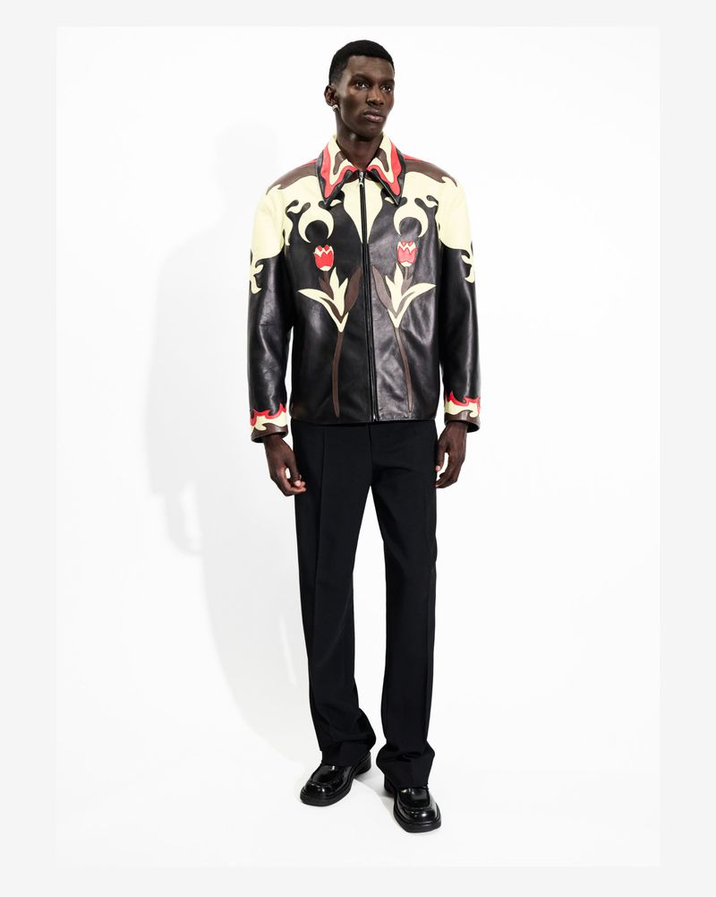 Full-length shot of a Black male model wearing a black leather jacket with floral accents, dark pants, and patent leather shoes.