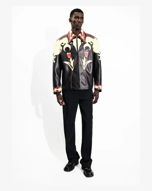 Full-length shot of a Black male model wearing a black leather jacket with floral accents, dark pants, and patent leather shoes.