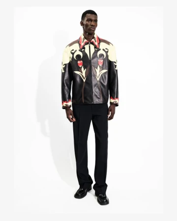 Full-length shot of a Black male model wearing a black leather jacket with floral accents, dark pants, and patent leather shoes.