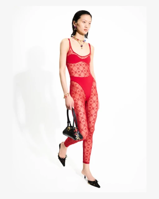 A slender Asian woman wearing a red bodysuit with a crescent moon pattern, jewelry, and black heels poses with a black handbag.