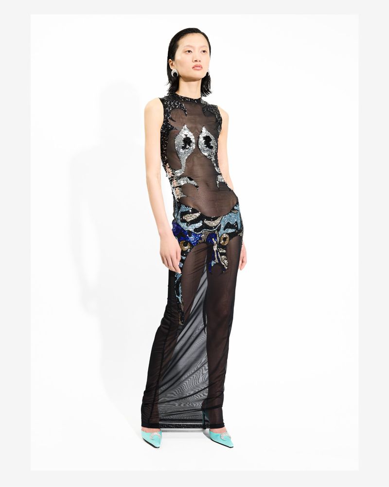 An Asian adult woman poses in a studio wearing a long, semi-sheer black dress with ornate silver, blue, and black embellishments. 