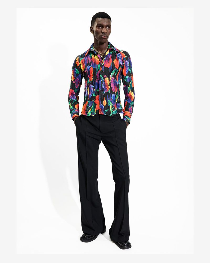 Full-length shot of a black male model in a floral shirt and black bell-bottom pants against a bright white seamless backdrop.