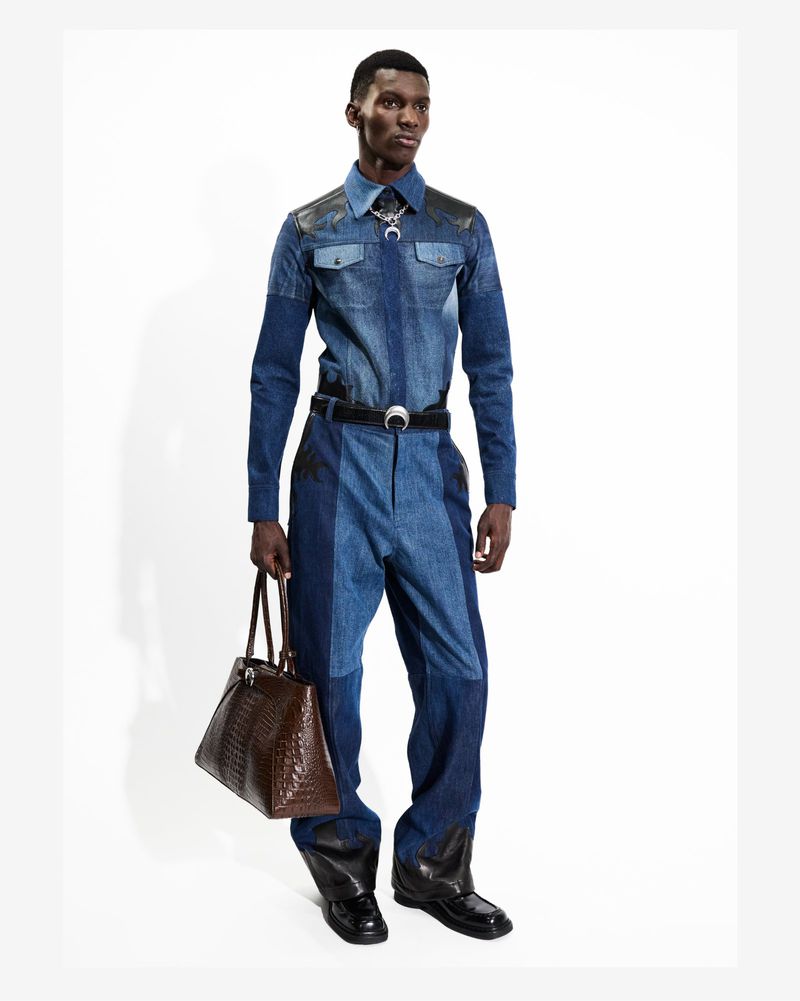 Fashion shot of a black male model in a denim jumpsuit, posing indoors with a brown leather bag against a white background.