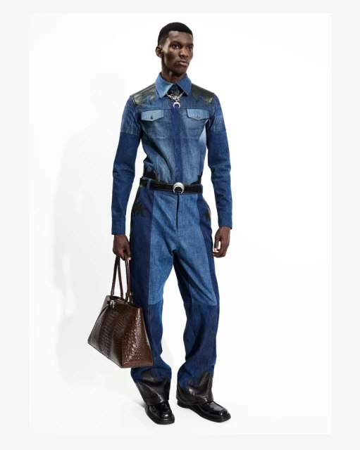 Fashion shot of a black male model in a denim jumpsuit, posing indoors with a brown leather bag against a white background.