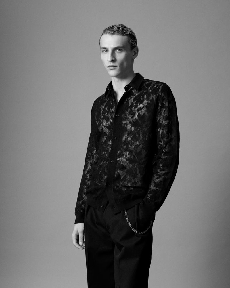 Black and white portrait of a young adult male fashion model wearing a black floral lace shirt and black pants against a grey background.