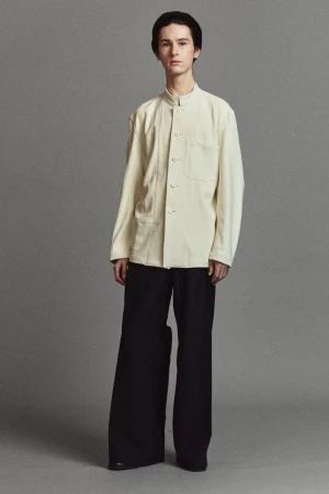 A young adult man posing in a studio wearing a light-colored, collared jacket and baggy black trousers, presented in a full studio shot.