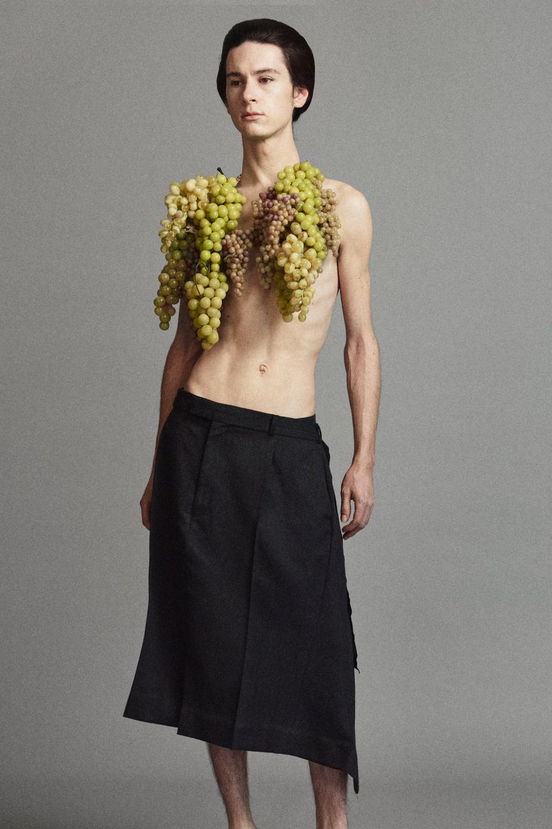 Male fashion model with dark hair poses topless in black skirt, with bunches of green grapes around his shoulders in a studio shot.