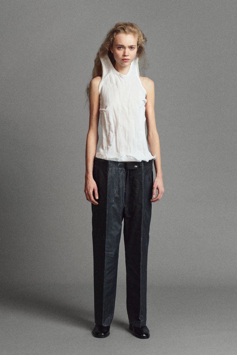 Model Ella Dalton in a crinkled white sleeveless top and dark tailored trousers posing against a grey studio backdrop.