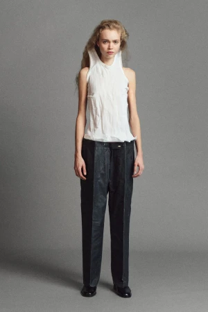 Model Ella Dalton in a crinkled white sleeveless top and dark tailored trousers posing against a grey studio backdrop.