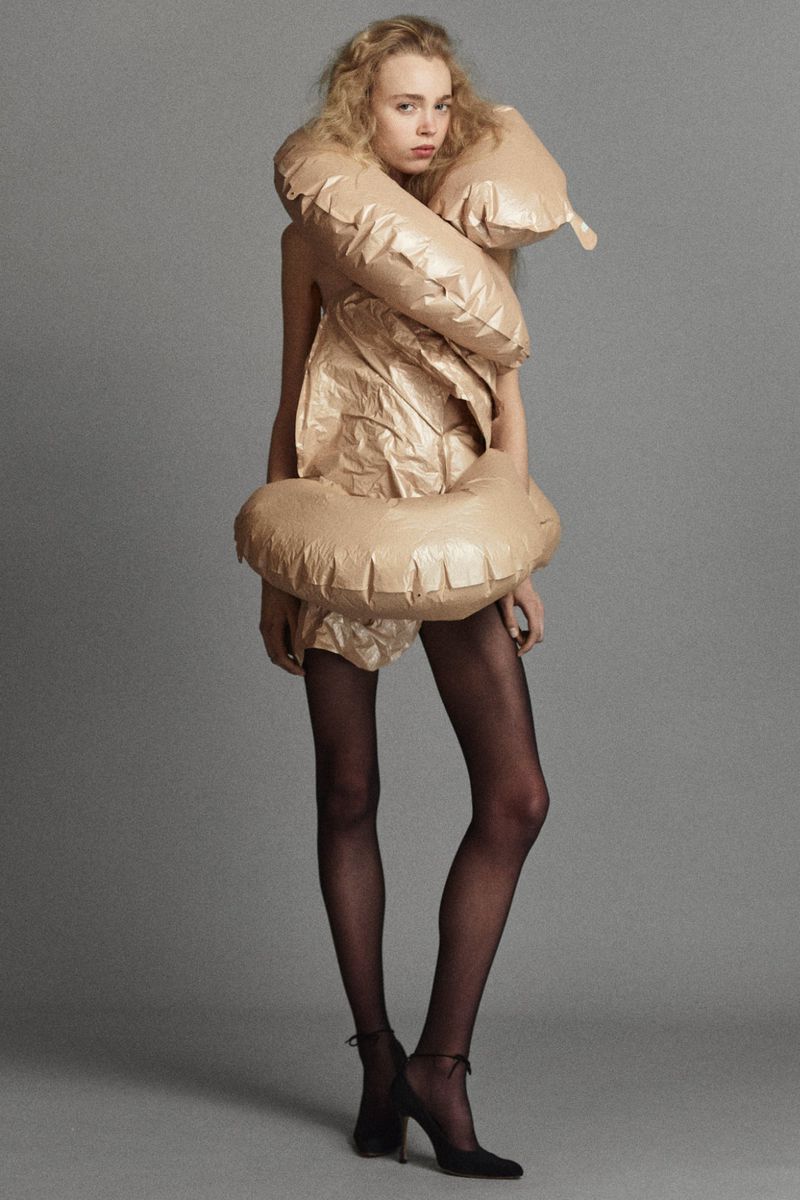 Model in an inflatable sculptural beige wrap garment with sheer black tights and black heels against a grey studio backdrop.