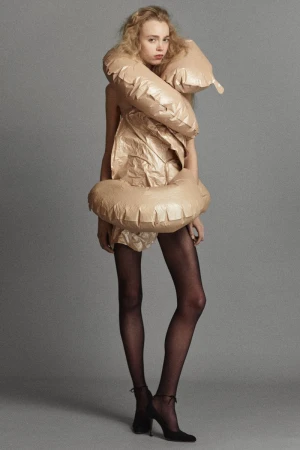 Model in an inflatable sculptural beige wrap garment with sheer black tights and black heels against a grey studio backdrop.