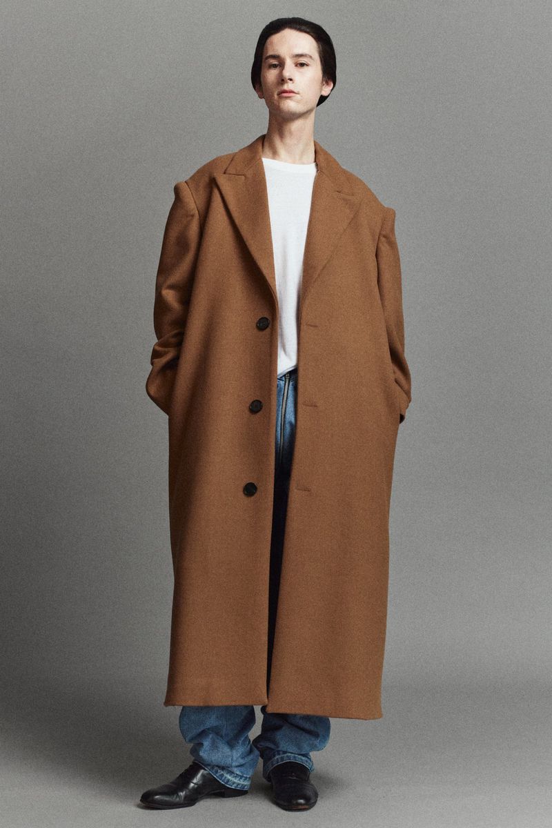 Model wearing an oversized camel brown wool overcoat with blue denim jeans and black shoes against a grey studio backdrop.