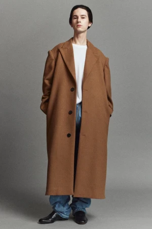Model wearing an oversized camel brown wool overcoat with blue denim jeans and black shoes against a grey studio backdrop.