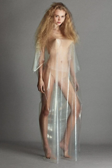 Woman in translucent plastic dress stands in a studio setting, showcasing modern fashion and minimalist aesthetic against a gray backdrop.